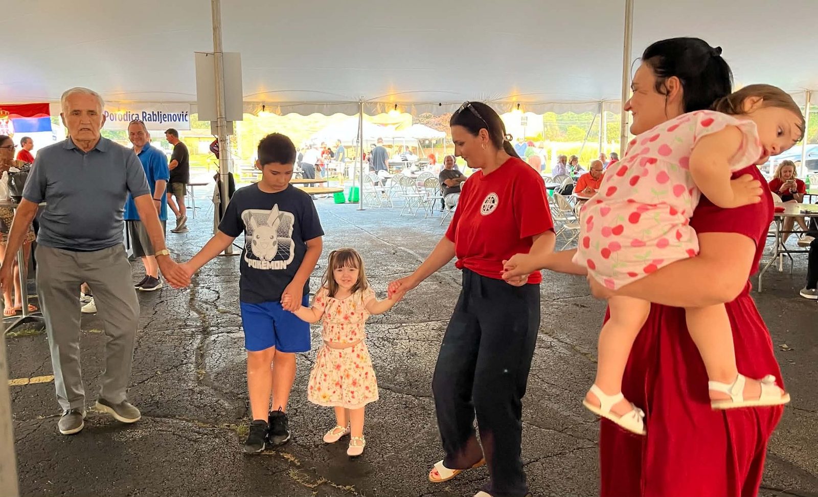 Three generations hold hands in the kolo, the traditional Serbian circle dance, under the festival tent at Serb Fest 2026